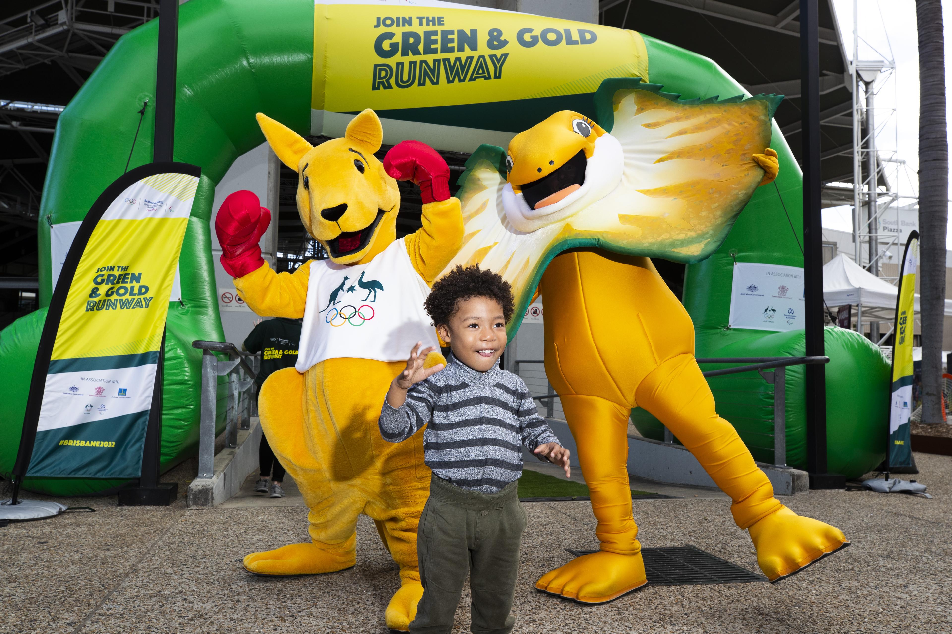 Boxing Kangaroo and Lizzie, a frill-necked lizard stand in front of a 'join the green and gold runway' blowup banner, with a young boy in front of them