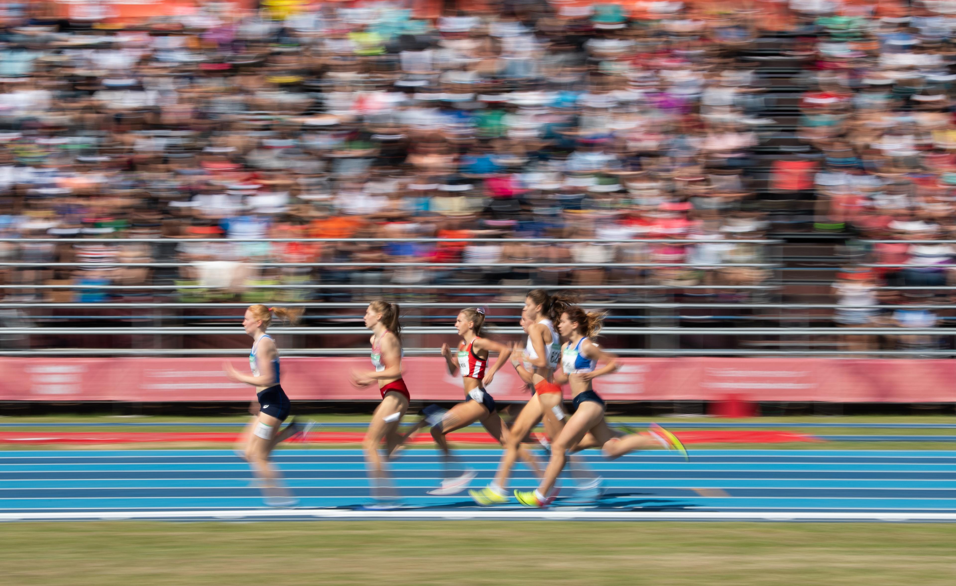 Female sprinters running on a blue athletics track with the crowd blurred in the background